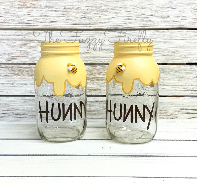 May include: Two clear glass jars with yellow paint and a dripping honey design. Each jar has a small bee figurine on the top and the word "Hunny" written in brown on the front.