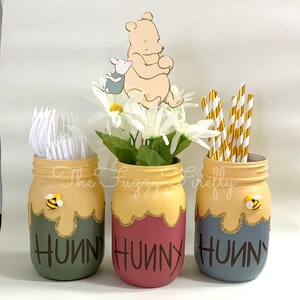 Peut inclure: Trois bocaux en verre peints utilis&eacute;s comme porte-ustensiles. Les bocaux pr&eacute;sentent une section sup&eacute;rieure jaune avec des gouttes de paillettes dor&eacute;es et le mot HUNNY en brun fonc&eacute;. Le pot de gauche est vert sauge, celui du centre est bordeaux.