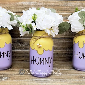 Peut inclure: Trois pots en verre peints en violet clair avec un motif de miel jaune qui coule. Chaque pot a un petit charme d'abeille et le mot "Hunny" &eacute;crit en noir. Les pots sont remplis de fleurs blanches.