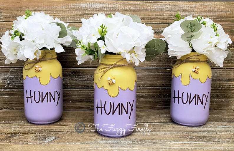 Peut inclure: Trois pots en verre peints en violet clair avec un motif de miel jaune qui coule. Chaque pot a un petit charme d'abeille et le mot "Hunny" &eacute;crit en noir. Les pots sont remplis de fleurs blanches.