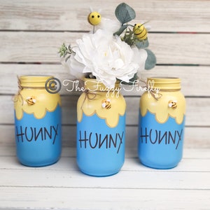 Peut inclure: Trois pots en verre bleus avec des couvercles jaunes et des nids d'abeilles peints dessus. Chaque pot porte le mot "Hunny" &eacute;crit en noir sur le devant. Les pots sont d&eacute;cor&eacute;s de petites figurines d'abeilles jaunes et noires.