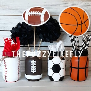 May include: Four mason jars decorated with sports themes. The jars are filled with red plastic forks, white plastic forks, black and white striped straws, and black and white straws. The jars are decorated with a basketball, a football, a baseball, and a soccer ball.
