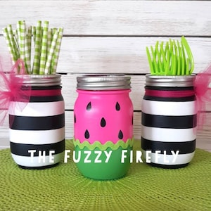 Puede incluir: Tres frascos de vidrio con tapas plateadas. Dos frascos tienen rayas horizontales blancas y negras con lazos de tul rosa. Un frasco est&aacute; pintado de rosa con semillas negras en forma de l&aacute;grima y una base verde. Texto THE FUZZY FIREFLY.