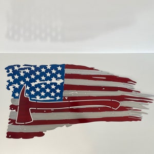 May include: A distressed American flag with a red, white, and blue color scheme. A silver firefighter axe is positioned across the flag, with the handle extending to the right.