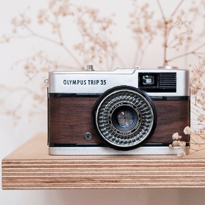 Op de afbeelding: Een vintage Olympus Trip 35 camera met een houtnerf afwerking. De camera is zilver en zwart met een zilveren lens. De camera staat op een houten plank.