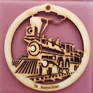 May include: A wooden ornament shaped like a circle with a cutout of a steam train engine. The ornament is light brown and has the words "St. Augustine" at the bottom.