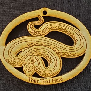 May include: A wooden ornament shaped like a circle with a snake design inside. The snake is curled up and has a detailed texture. The ornament has a space for text at the bottom that says "Your Text Here".