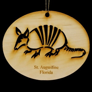 May include: A wooden ornament shaped like an armadillo with a black silhouette. The ornament has the text "St. Augustine Florida" on the bottom.