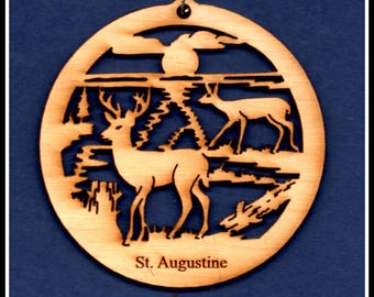 May include: A wooden Christmas ornament featuring a silhouette of two deer in a forest setting. The ornament is round and has a brown wood finish. The text "St. Augustine" is printed on the bottom of the ornament.
