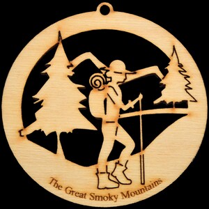 May include: A wooden ornament featuring a silhouette of a hiker with a backpack and walking stick, standing in front of two pine trees. The ornament is inscribed with the text "The Great Smoky Mountains".