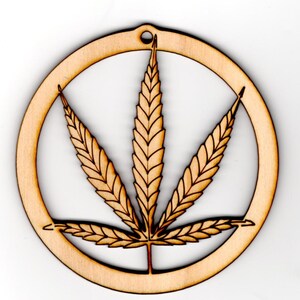 May include: A wooden ornament in the shape of a circle with a cutout of a marijuana leaf. The ornament is light brown in color.
