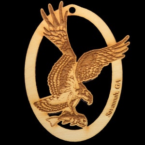 May include: A wooden ornament shaped like an oval with a hawk or falcon in flight, holding a fish in its talons. The ornament is laser-cut and engraved with the words "Savannah, GA".