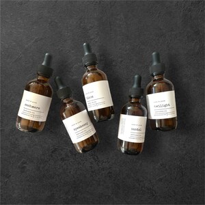 May include: Five brown glass diffuser oil bottles with black droppers and white labels. The labels read "Cashmere", "Bloom", "Speakeasy", "Santal", and "Twilight". The bottles are arranged on a dark grey surface.