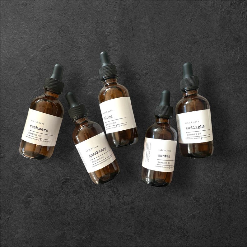May include: Five brown glass diffuser oil bottles with black droppers and white labels. The labels read "Cashmere", "Bloom", "Speakeasy", "Santal", and "Twilight". The bottles are arranged on a dark grey surface.