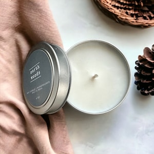 May include: A silver tin candle with the text "north woods" on the label. The candle is hand-poured and contains salt and peak. The candle is 4 oz. The candle is next to a pink cloth, a pine cone, and a woven basket.