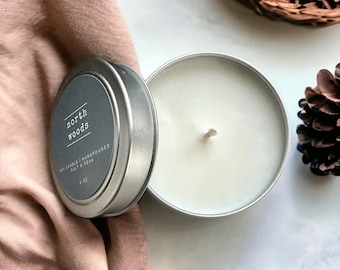 May include: A silver tin candle with the text "north woods" on the label. The candle is hand-poured and contains salt and peak. The candle is 4 oz. The candle is next to a pink cloth, a pine cone, and a woven basket.