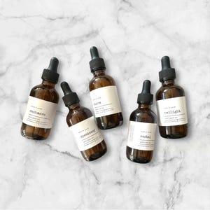 May include: Five amber glass diffuser oil bottles with black droppers. Each bottle has a white label with a scent name: Cashmere, Speakeasy, Bloom, Santal, and Twilight. The bottles are arranged on a white and grey marble surface.