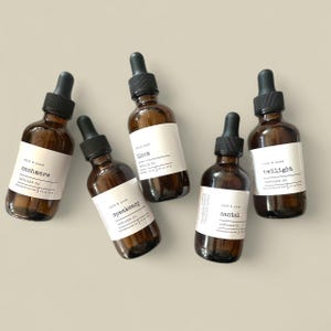 May include: Five brown glass diffuser oil bottles with black droppers and white labels. The labels read "cashmere", "bloom", "speakeasy", "santal", and "twilight". The bottles are arranged on a light beige surface.