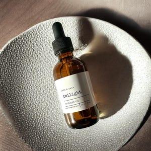 May include: A brown glass bottle with a black dropper, labelled "twilight diffuser oil." The label also includes the text "salt & peak" and "2 fl oz | 59 ml." The bottle sits on a textured, white, decorative plate.