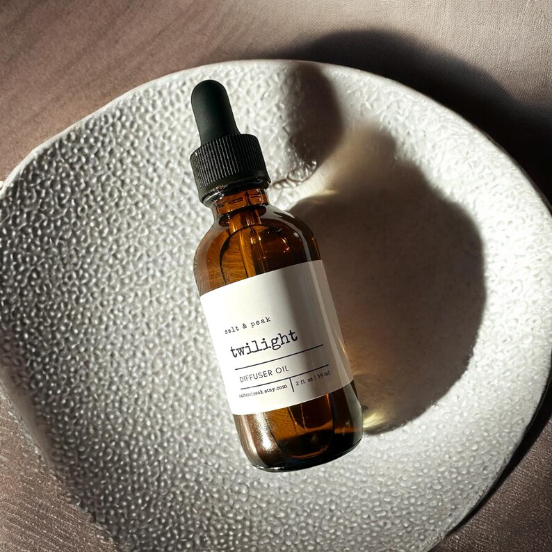 May include: A brown glass bottle with a black dropper, labelled "twilight diffuser oil." The label also includes the text "salt & peak" and "2 fl oz | 59 ml." The bottle sits on a textured, white, decorative plate.