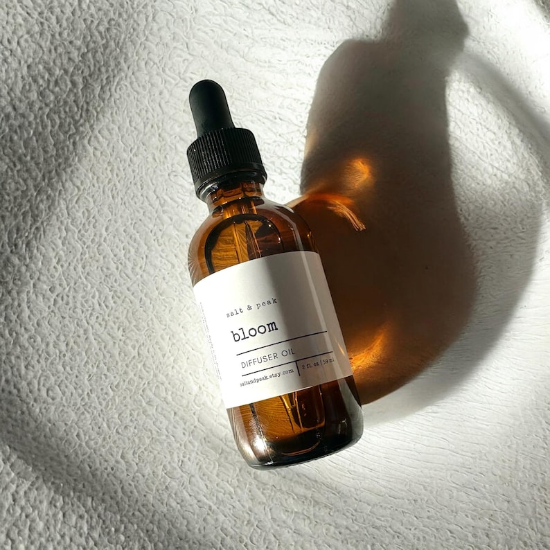 May include: A brown glass diffuser oil bottle with a black dropper. The label reads "salt & peak bloom DIFFUSER OIL". The bottle is 59 ml. The bottle is on a white textured surface.