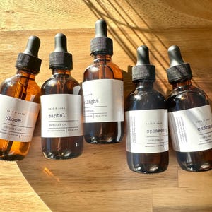 May include: Five amber glass diffuser oil bottles with black droppers and white labels. The labels read "bloom", "santal", "starlight", "speakeasy", and "cashmere". The bottles are arranged on a wooden surface.