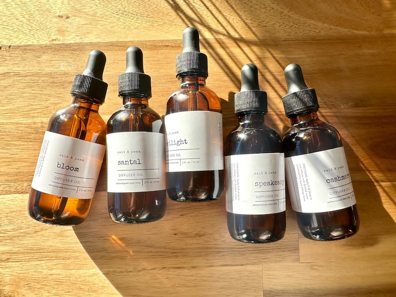 May include: Five amber glass diffuser oil bottles with black droppers and white labels. The labels read "bloom", "santal", "starlight", "speakeasy", and "cashmere". The bottles are arranged on a wooden surface.