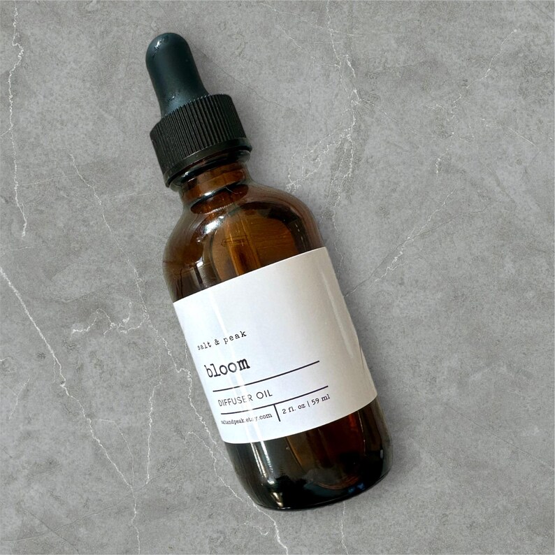 May include: A brown glass bottle with a black dropper top and a white label. The label reads "bloom DIFFUSER OIL 2 fl oz | 59 ml" and "salt & peak". The bottle is angled on a grey surface.