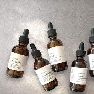May include: A collection of amber glass diffuser oil bottles with black droppers, arranged on a grey surface. Each bottle features a white label with the words "cashmere", "bloom", "speakeasy", and "santal". The bottles are labeled "DIFFUSER OIL".