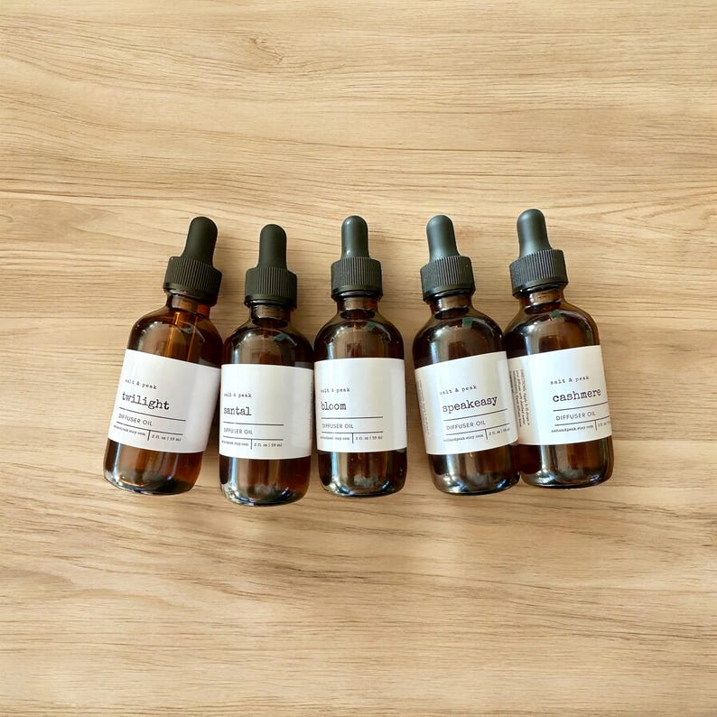 May include: Five amber glass bottles with black droppers and white labels. The labels read "twilight", "santal", "bloom", "speakeasy", and "cashmere". Each bottle contains diffuser oil. The bottles are arranged on a light brown wooden surface.