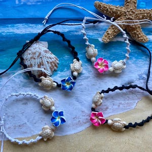 May include: Four adjustable bracelets featuring woven cords in black or white. Each bracelet includes two small cream-colored turtle charms and a single five-petaled flower charm in either vibrant blue with a yellow center or bright pink with a white center.