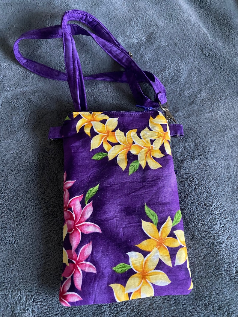 May include: A purple crossbody bag with a floral pattern. The bag features yellow and pink flowers with green leaves. It has a zipper closure and a purple strap.