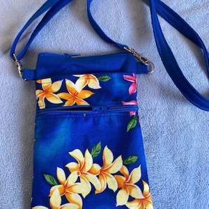 May include: A blue crossbody bag with a floral pattern featuring yellow and orange flowers and green leaves. The bag has a blue adjustable strap and a zippered main compartment. The bag is made of fabric.