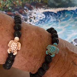 May include: Two black lava stone bracelets, each featuring a turtle charm. One turtle is off-white, the other is turquoise. The bracelets are a dark, earthy accessory.