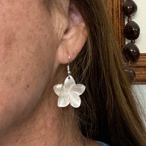 May include: A pair of flower-shaped earrings. Each earring features a five-petal design with a pearlescent, iridescent finish. The earrings are suspended from silver-colored hooks. The earrings are a jewelry item.