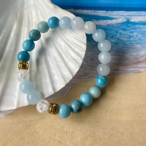 May include: A beaded bracelet featuring various shades of blue and white gemstones. The bracelet includes gold-colored spacers and is displayed on a seashell against a beach scene background. The beads vary in size and texture.