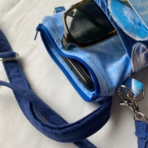May include: A blue fabric crossbody bag with a zippered compartment, holding sunglasses, a phone, and a twenty-dollar bill. The bag has a blue strap and a silver clasp. The bag is patterned with blue and white designs.