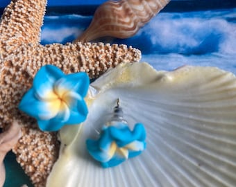 Può includere: Un paio di orecchini a bottone a forma di fiore blu e giallo. Gli orecchini sono esposti su una conchiglia bianca, con una stella marina e una conchiglia sullo sfondo. Lo sfondo presenta una scena oceanica blu.