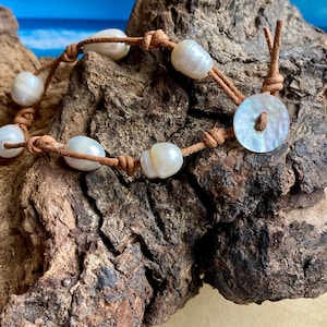 May include: A handmade bracelet featuring lustrous, off-white pearls strung on brown leather cord. The bracelet has a button closure made of a mother-of-pearl disc. The background shows a blurred image of the ocean and sky.