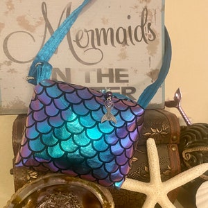 May include: A small, square purse with a mermaid scale pattern in shades of blue and purple. The purse has a blue strap and a silver mermaid tail charm. The background includes a sign that says "Mermaids" and decorative seashells.