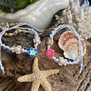 May include: White macrame bracelets with turtle and flower charms. A starfish and seashell are displayed on a piece of driftwood. The bracelets have a tropical theme.