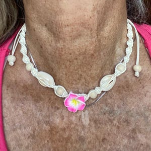 May include: A white macrame necklace featuring seashells, wooden beads, and a pink and yellow flower pendant. The necklace is adjustable with a sliding knot closure. The necklace is a beach-themed accessory.