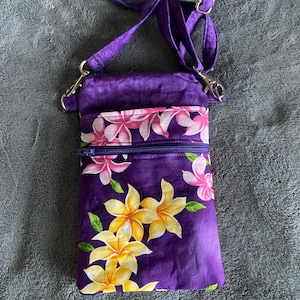 May include: A purple crossbody bag with a floral pattern. The bag features a zipper closure and an adjustable strap. The floral design includes yellow and pink flowers with green leaves. The bag is made of fabric.