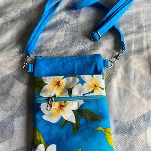 May include: A blue crossbody bag with a floral pattern of white and yellow flowers. The bag has a zippered front pocket and a blue adjustable strap. The bag is made of fabric and has silver-colored metal hardware.