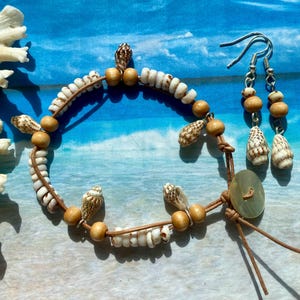 May include: A handmade bracelet and matching earrings set. The bracelet features small white shells, wooden beads, and small seashells strung on a brown cord. The earrings have wooden beads and shell pendants. The set is on a blue and white background.