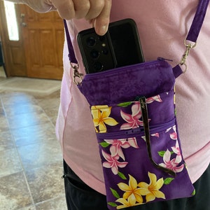 May include: Purple crossbody phone bag with a floral pattern of yellow and pink flowers. The bag has a zippered pocket and a strap. A black phone is in the top pocket, and sunglasses are in the front pocket.