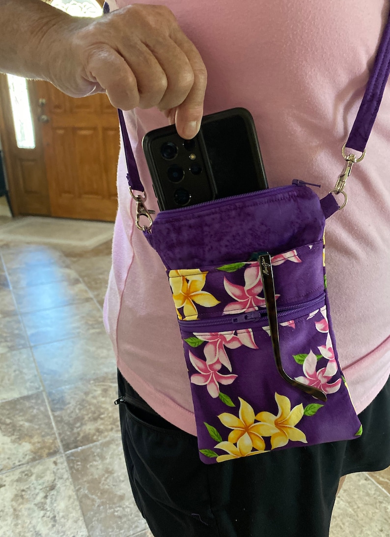 May include: Purple crossbody phone bag with a floral pattern of yellow and pink flowers. The bag has a zippered pocket and a strap. A black phone is in the top pocket, and sunglasses are in the front pocket.