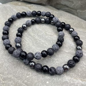 May include: Two beaded bracelets. One bracelet features black, gray, and silver beads. The other bracelet is made of black and gray lava rock beads. The bracelets are displayed on a gray stone surface.