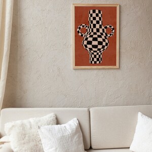 May include: A framed print of a black and white chequered vase with a red background. The vase has two handles and a rounded body.