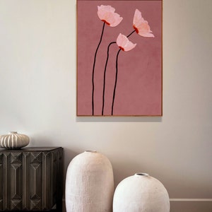 May include: A framed floral art print featuring three pink flowers with black stems against a dusty rose background. The artwork is in a gold frame and is displayed on a white wall. The style is minimalist and modern.